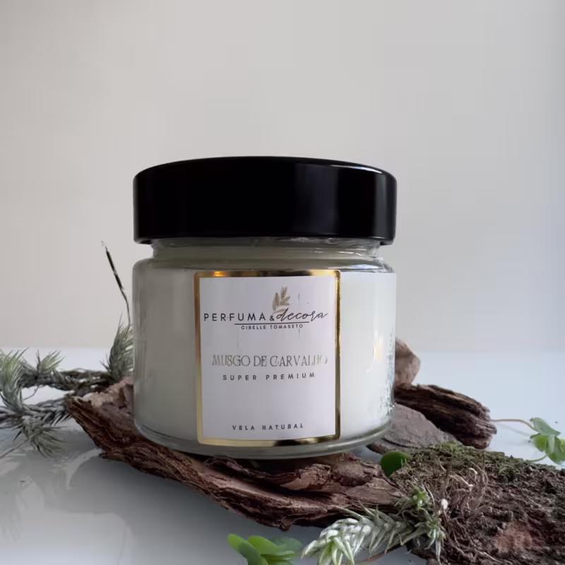 Frasco de vidro transparente com vela e tampa preta. Rótulo na frente com o nome "Perfuma & Decora" e "Musgo de Carvalho Super Premium". O frasco está posicionado sobre pedaços de casca de árvore e ramos com folhas verdes.