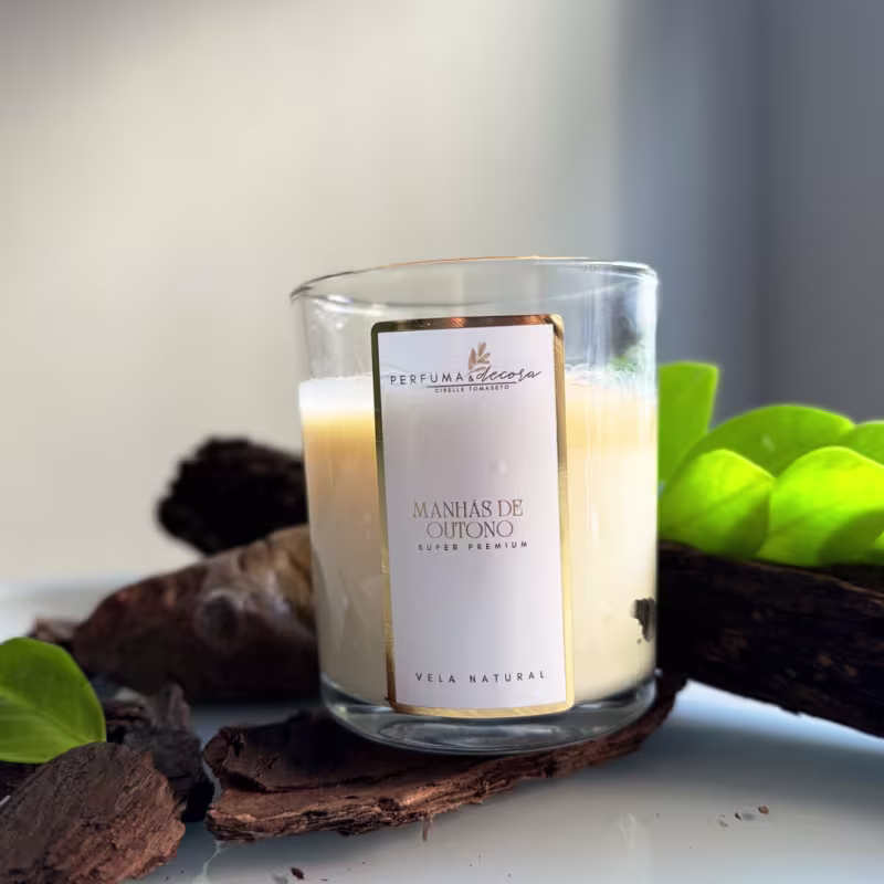 Vela natural em frasco de vidro transparente. O frasco tem uma etiqueta branca com detalhes em dourado que diz "Perfuma & Decora Manhãs de Outono". Ao redor do frasco, há folhas verdes e pedaços de casca de árvore, criando um ambiente outonal.