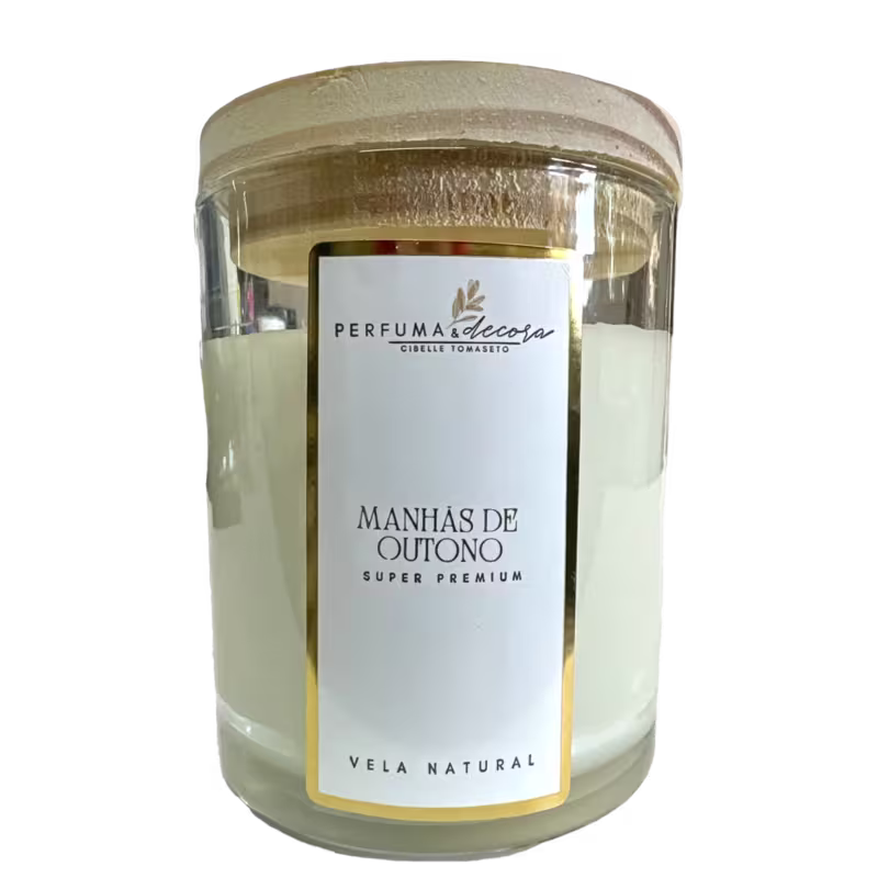Vela em vidro transparente com tampa de madeira. A embalagem tem um rótulo branco com o nome do produto "Manhãs de Outono" da marca "Perfuma & Decora". O frasco tem uma forma cilíndrica e as cores predominantes são branco, dourado e transparente.