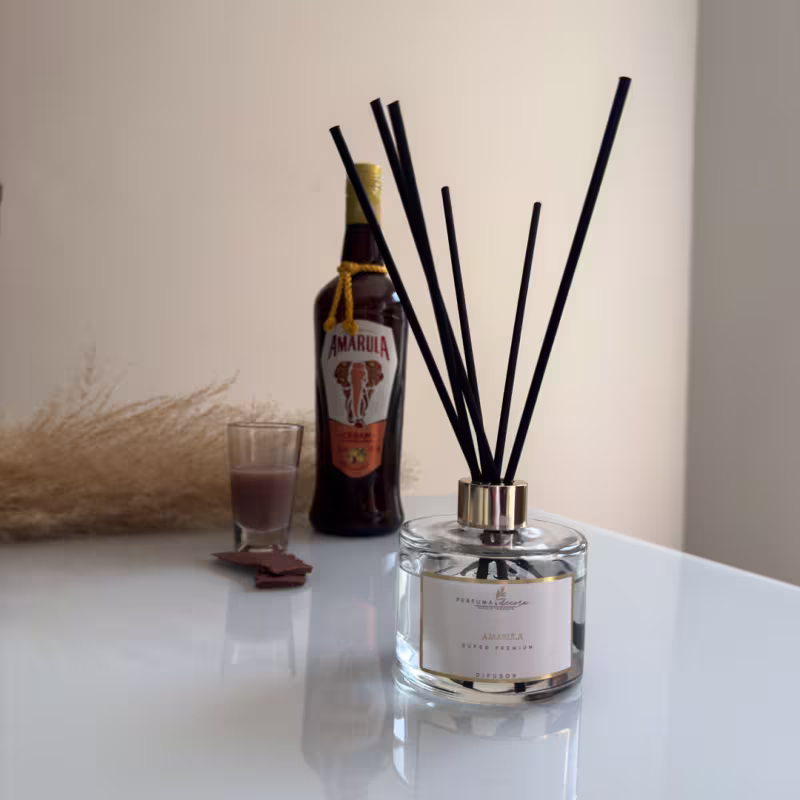 Difusor de aromas em frasco de vidro transparente com uma tampa metálica. Dentro do frasco, há um conjunto de hastes pretas que servem para difundir o aroma. Ao lado do difusor, há uma garrafa de licor Amarula.