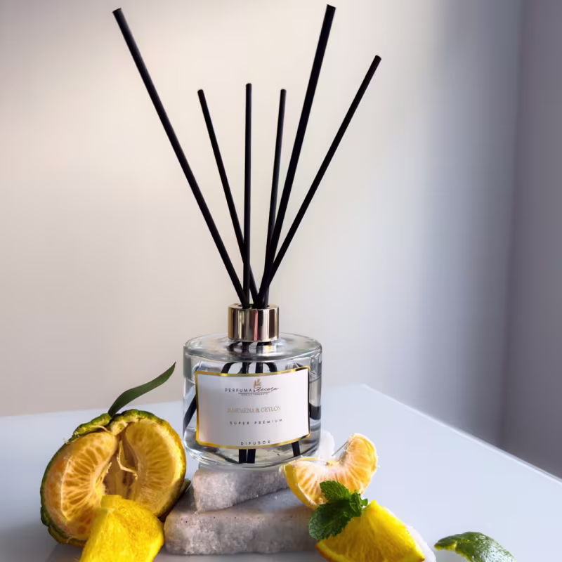 Frasco de vidro transparente com tampa dourada. Contém hastes pretas para difusão do aroma.  Ao redor do frasco, há fatias de laranja, folhas verdes e pedras decorativas. As cores predominantes são amarelo, verde, branco e preto.