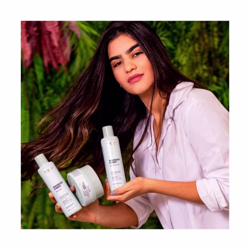 A imagem mostra uma mulher segurando 3 produtos da linha Proboo Fiber da marca Prohall Cosmetic.