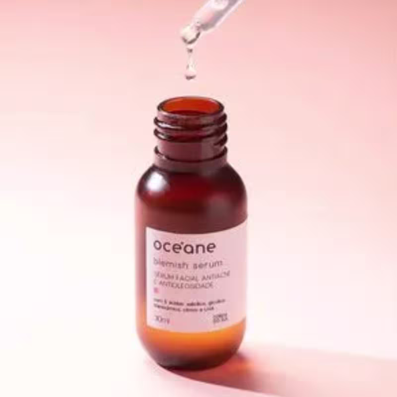A imagem mostra o frasco do  "Océane Blemish Serum - Sérum Facial Antiacne e Antioleosidade" aberto, acima dele o conta-gotas está pingando o líquido transparente. Tudo isso está sobre um fundo rosado.