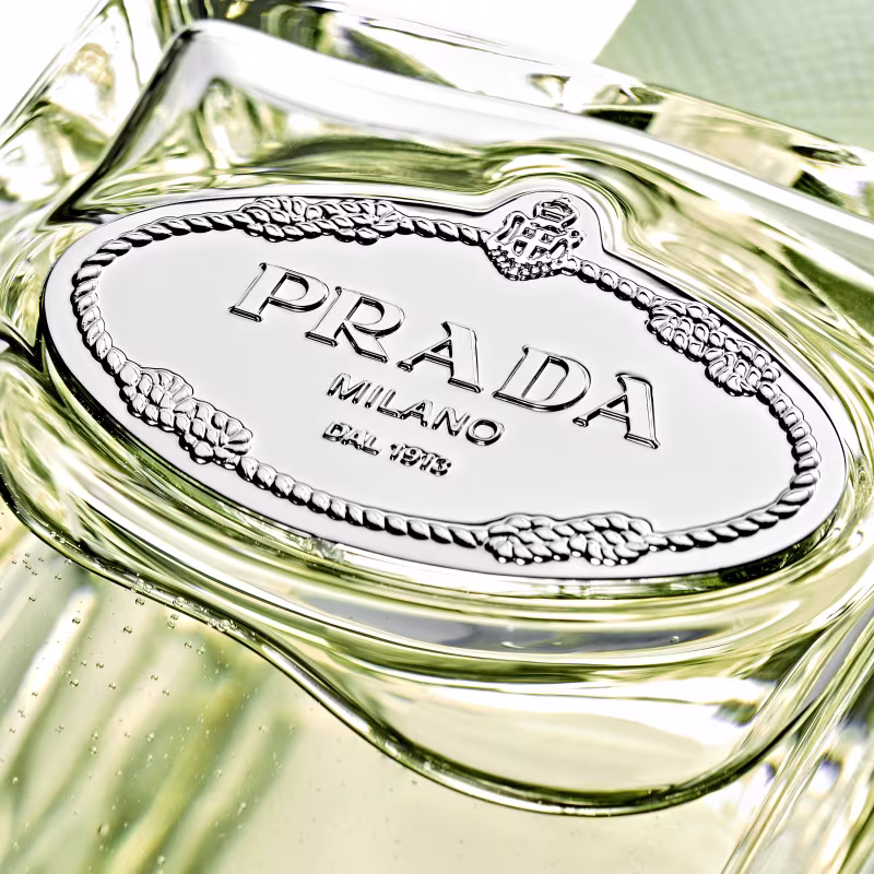 Perfume em frasco de vidro transparente com detalhes prateados. O frasco tem formato retangular com cantos arredondados. O rótulo oval prateado no corpo do frasco exibe o nome "PRADA" em letras maiúsculas, seguido de "MILANO" e o número "DAL 1913".