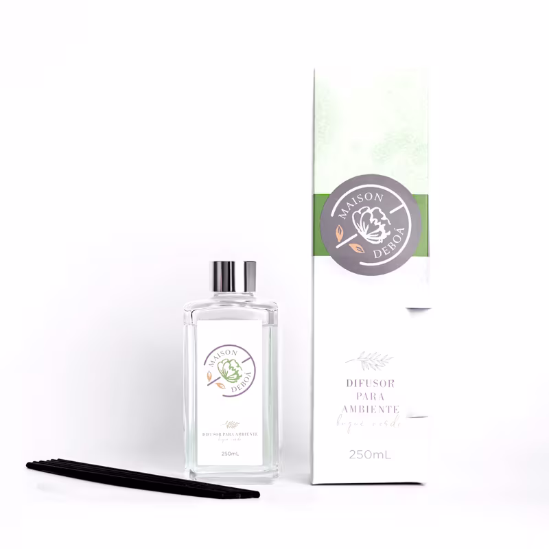 Difusor de ambientes em frasco transparente de vidro com tampa prateada. Embalagem retangular branca com detalhes em verde, exibindo o logotipo "Maison Deboá". Bastões pretos e caixa retangular acompanha.