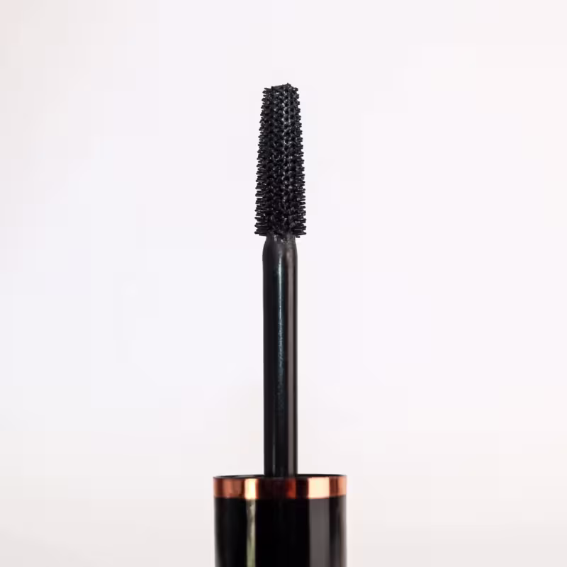 Rímel em formato de tubo preto com detalhes em cobre na base. O aplicador possui cerdas densas e compridas na cor preta. O produto é da marca Ustē Cosméticos.