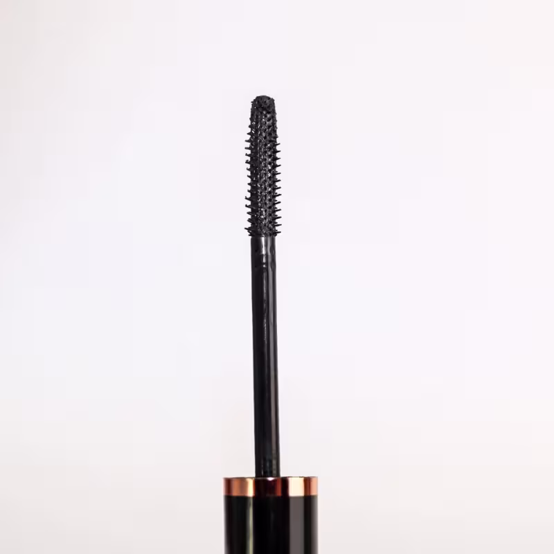 Rímel em tubo preto com detalhes em cobre na base. O aplicador é um pincel cilíndrico com cerdas pretas projetado para definir e volumizar os cílios. O produto tem uma aparência sofisticada e elegante, adequado para criar um olhar marcante e impactante.