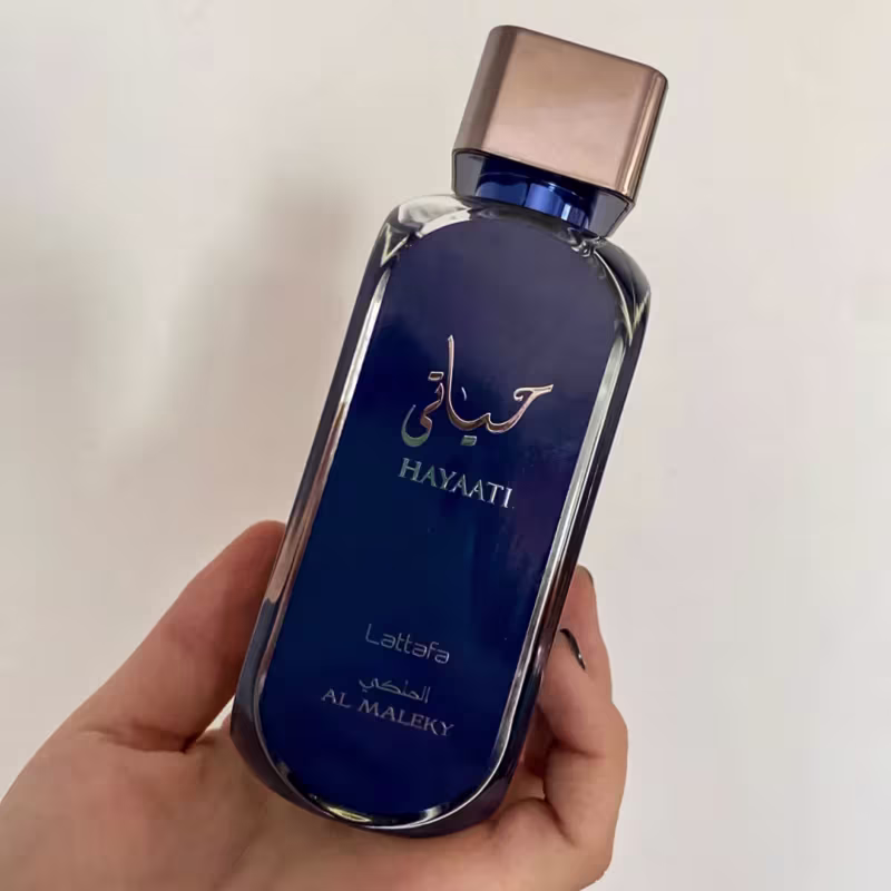 A imagem mostra o perfume sendo segurado por uma mão feminina.