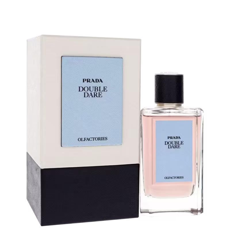 Perfume em frasco retangular transparente com líquido rosa pálido. Rótulo azul claro com o texto "PRADA DOUBLE DARE" e "OLFACTORIES". Tampa preta texturizada. Embalagem minimalista e elegante, típica da marca Prada.