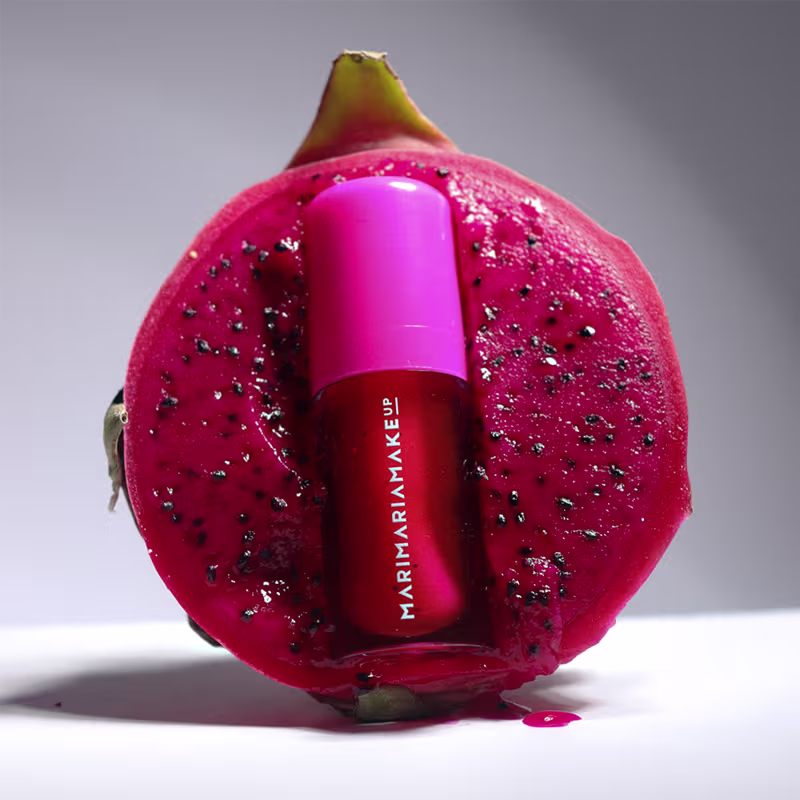 A imagem mostra o Lip Oil da marca Mari Maria Makeup, destacado em uma pitaya, criando um contraste de cores vibrantes.