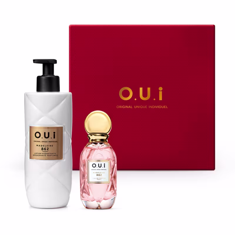 A imagem mostra um frasco transparente com eau de parfum rosa com detalhes dourados e a inscrição "O.U.i" em letras grandes. Ao lado, há um vidro plástico branco com a tampa preta que traz um hidratante corporal e ao fundo uma caixa de presente vermelha.
