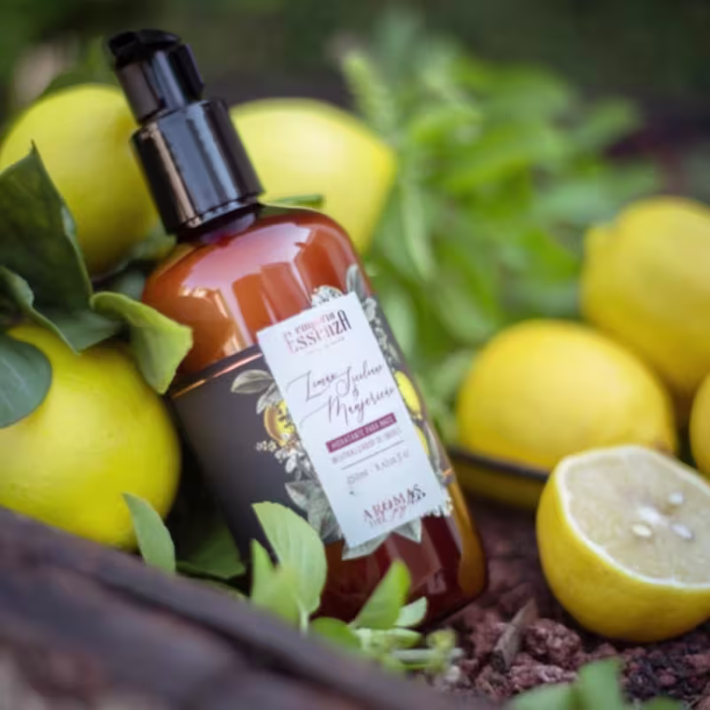 Frasco do hidratante de mãos Empório Essenza Serra Gaúcha, em embalagem marrom com tampa preta, cercado por limões amarelos e folhas verdes.