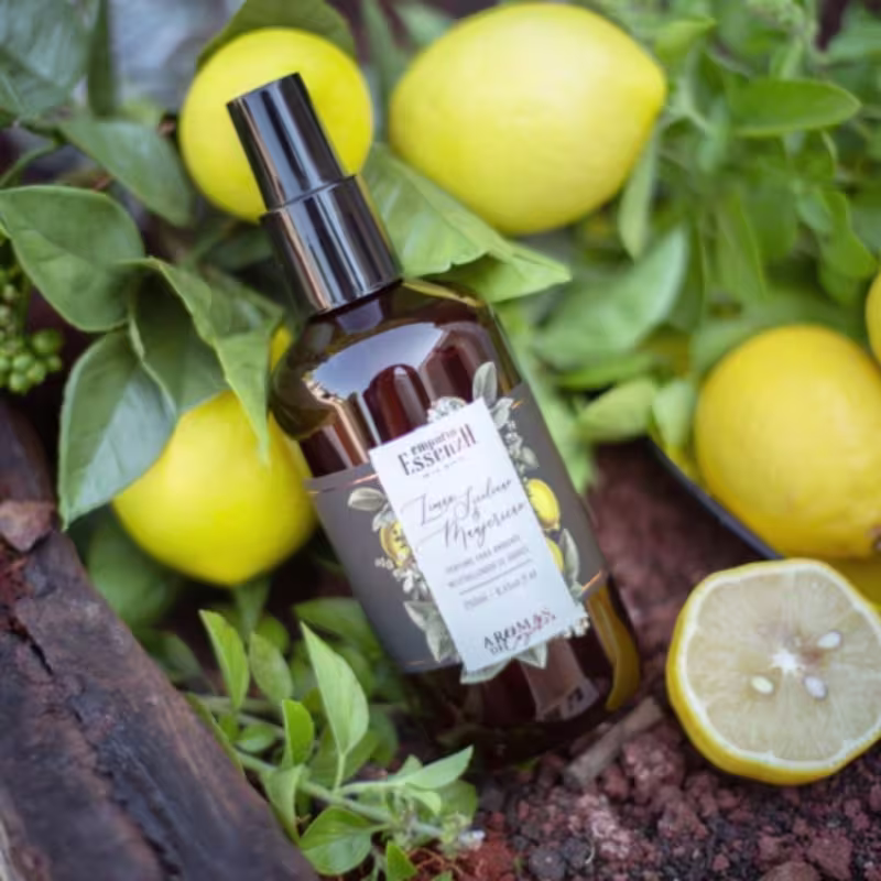 Frasco marrom com rótulo da marca Empório Essenza Serra Gaúcha, contendo perfume de ambiente com aroma de limão, cercado por folhas verdes e limões amarelos.