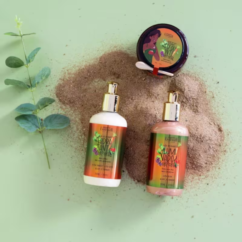 Ambiente com produtos da marca Empório Essenza Serra Gaúcha, com embalagens em tons de verde e laranja, sobre superfície arenosa e folhagem verde, sugerindo um contexto natural e orgânico.