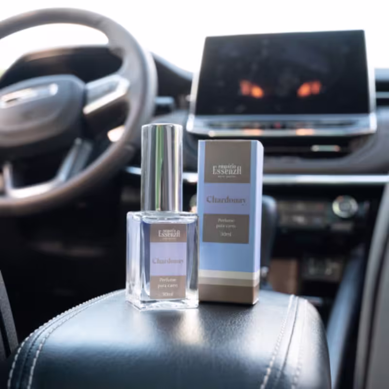 Frasco de perfume Essenza Serra Gaúcha da linha Chardonnay, com embalagem cinza e branca, exibindo o nome do produto em destaque. Ao fundo, painel de controle de veículo.