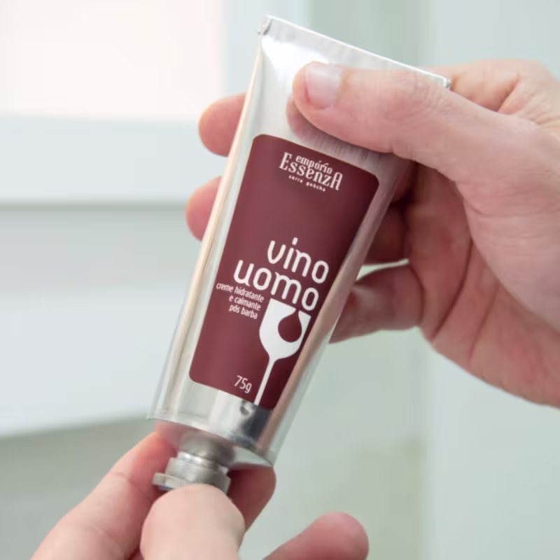 A imagem mostra um tubo de creme da marca Empório Essenza Serra Gaúcha, com o nome "Vino Uomo" destacado. O produto é segurado por uma mão, em um ambiente claro e minimalista, sugerindo um contexto de cuidados pessoais.