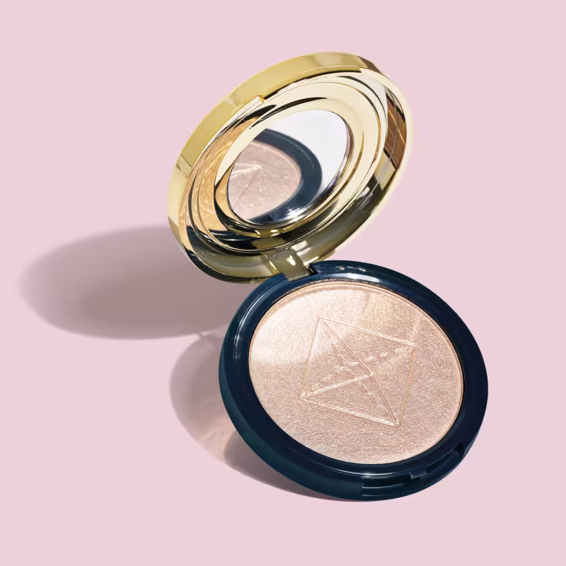 Produto de maquiagem em pó compacto com acabamento iluminado. Embalagem redonda azul com tampa dourada espelhada em camadas concêntricas. O pó compacto apresenta um acabamento perolado dourado. O design da embalagem dourado luxuoso contrasta com o azul.