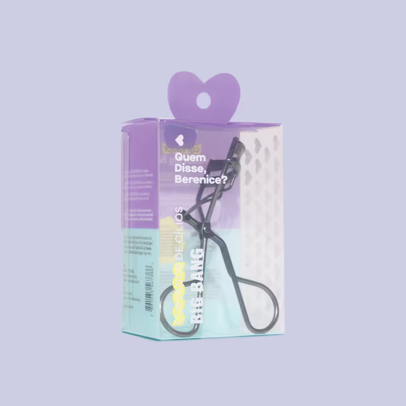 A imagem mostra uma embalagem plástica transparente que traz o  curvador de  cílios. A embalagem tem uma paleta de cores que vai do roxo no topo até um verde claro na parte inferior.