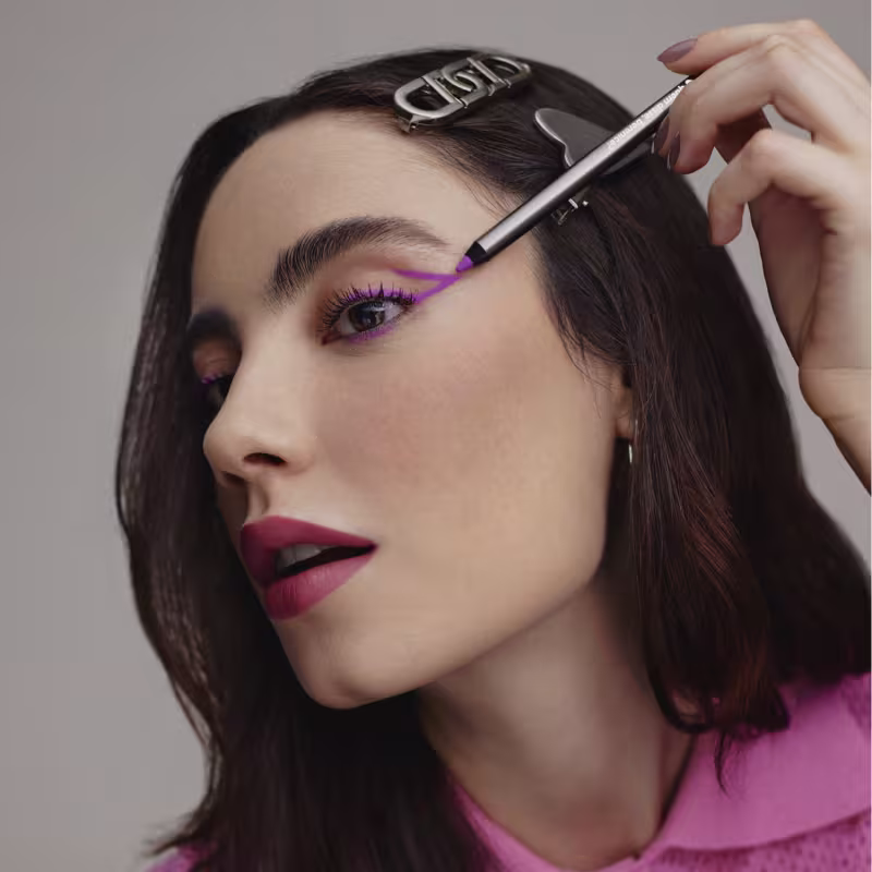 A imagem mostra uma modelo usando maquiagem de olhos em tons de roxo vibrante. Ela tem sobrancelhas bem delineadas e batom vermelho escuro nos lábios. Seu cabelo castanho escuro está penteado para trás, permitindo ver claramente seu rosto.