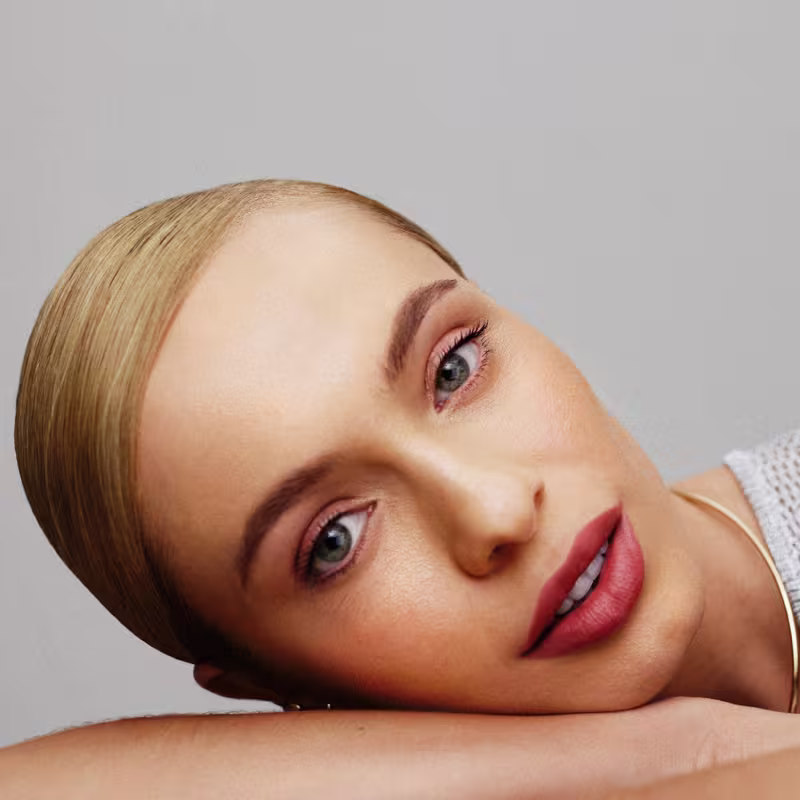 A imagem é um rosto feminino com maquiagem. Observa-se uma pele branca, olhos azuis expressivos, sobrancelhas bem definidas e lábios com um tom de rosa suave. A iluminação e o fundo neutro destacam as características faciais de forma elegante e sutil.