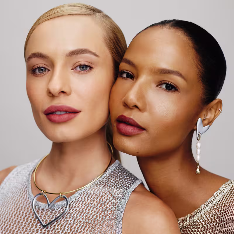 A imagem mostra dois rostos femininos em close-up, com expressões sérias e olhares diretos. Ambas as mulheres usam maquiagem com tons neutros e naturais, destacando seus traços faciais. Elas vestem roupas com detalhes em malha metálica.