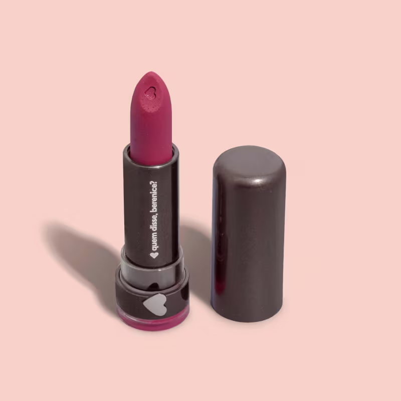 Barra de batom rosa escuro em embalagem preta com logotipo em branco. Produto de maquiagem com formato cilíndrico e tampa protetora.