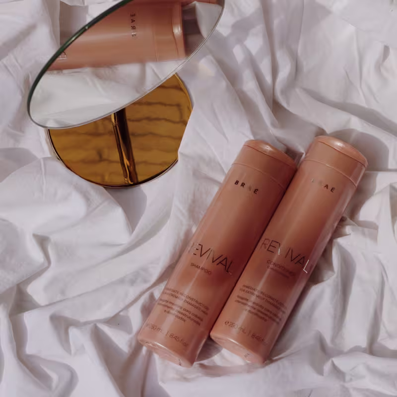 A imagem mostra shampoo e condicionador Revival da Braé. Eles estão deitados e ao lado dos frascos, há um espelho de maquiagem redondo com moldura dourada. Os itens estão dispostos sobre um tecido branco amarrotado.