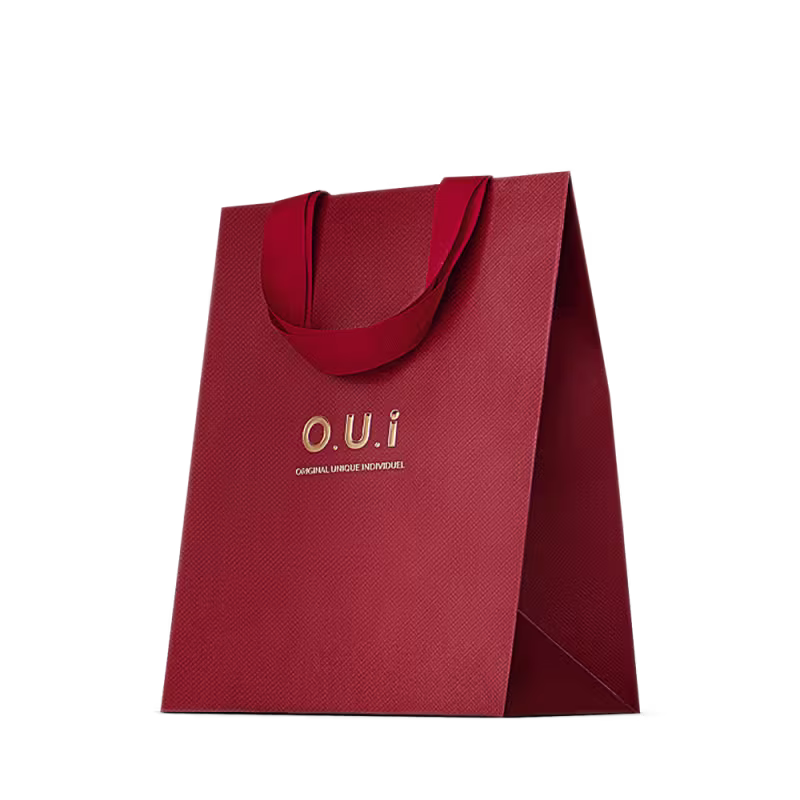 Sacola de presente vermelha em material texturizado, com alças e detalhes na mesma cor. Apresenta o logotipo "O.U.i" em letras douradas. A embalagem tem um design minimalista e elegante, com o vermelho vibrante como cor predominante.