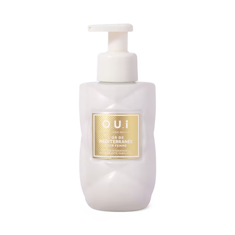 É um frasco plástico branco opaco com bomba dosadora para loção. O frasco tem uma etiqueta dourada quadrada com detalhes em relevo, exibindo o texto "OUI FLOR DE MEDITERRANEE " em letras maiúsculas e minúsculas.