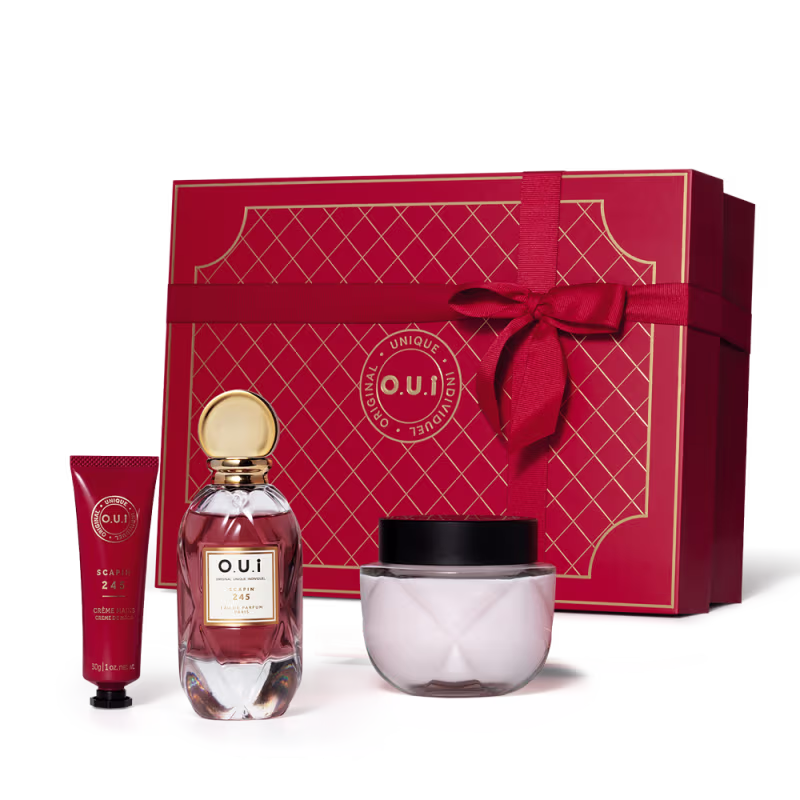 Conjunto. A peça central é o frasco do perfume em vidro transparente com líquido rosa claro e detalhes dourados na tampa e rótulo. Acompanha creme em embalagem redonda de vidro transparente, um frasco tubo vermelho de creme para mãos e caixa de presente