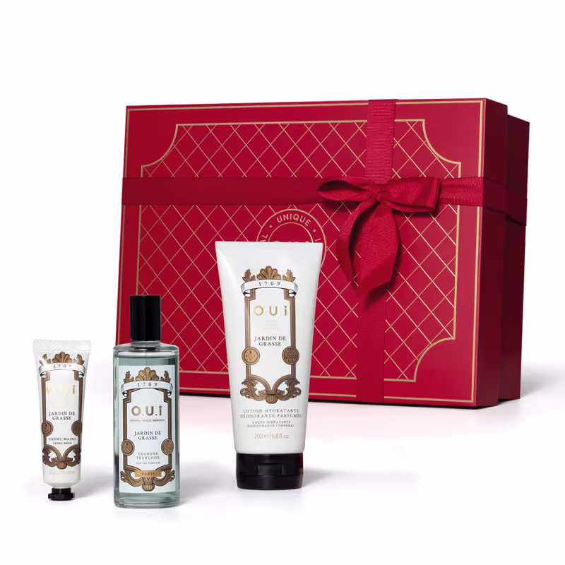 A imagem mostra um conjunto de produtos O.U.i em frente a uma bela caixa de presente vermelha. Ela traz um frasco com eau de parfum e dois tubos brancos com tampa preta, um menor com hidratante para as mãos e outro maior com hidratante para o corpo.