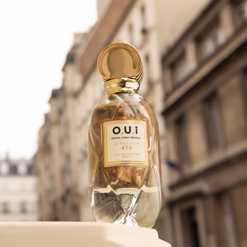 Frasco de perfume da marca OUI, modelo "La Villette 470", em primeiro plano, com fundo desfocado de uma construção arquitetônica. O produto é apresentado em um ambiente urbano, sugerindo um contexto elegante e sofisticado.