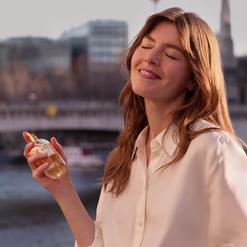 A mulher segura um frasco de perfume em um cenário urbano com edifícios ao fundo, sugerindo um ambiente moderno e sofisticado para o uso do produto de beleza.