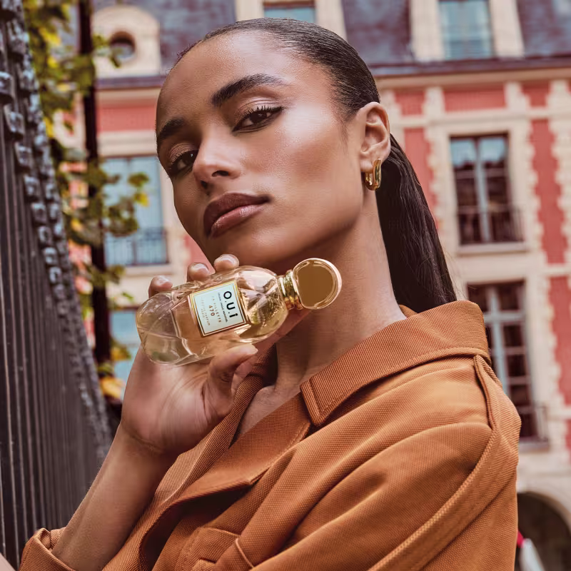 A mulher segura um frasco de perfume da marca Oui em uma rua urbana com edifícios históricos ao fundo, vestindo um casaco laranja, sugerindo um ambiente sofisticado e elegante para o uso do produto.