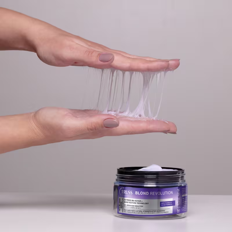 Duas mãos demonstrando o efeito teia que a máscara capilar possui sobre um pote roxo da marca Truss Blond Revolution, um produto de tratamento para cabelos loiros.