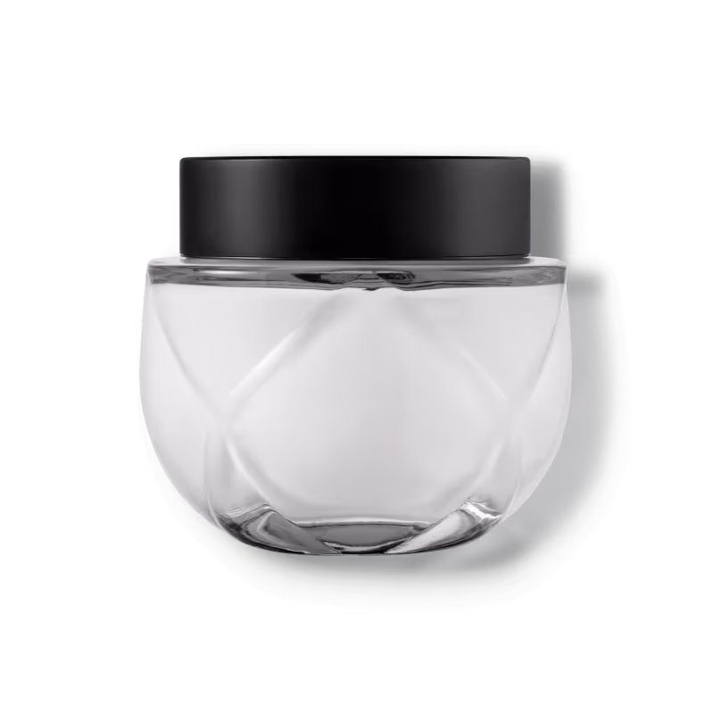 Frasco de vidro transparente arredondado com tampa preta. O recipiente tem uma forma esférica achatada. A tampa preta opaca contrasta com o vidro transparente do frasco que contém o creme na cor branca.