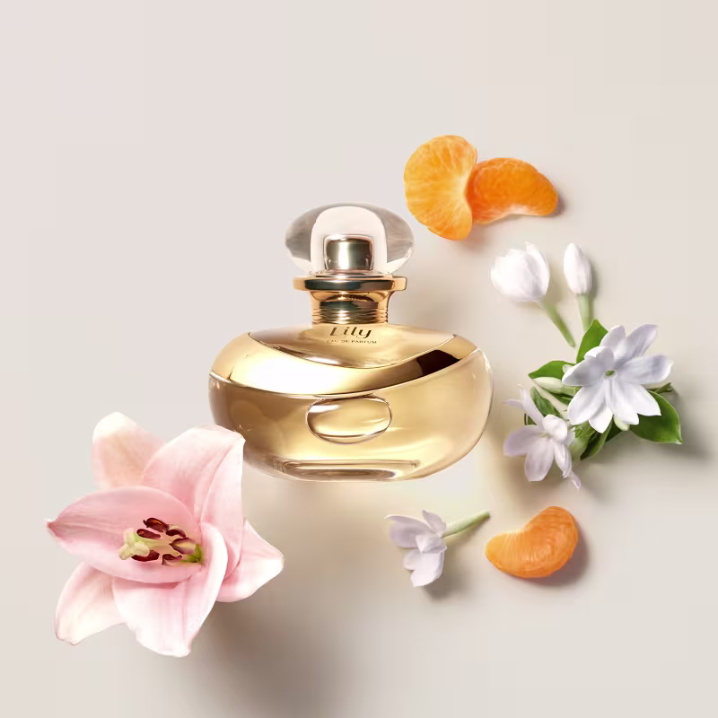A imagem apresenta um frasco de perfume dourado da marca Lily. Ao redor do frasco, há decorações florais delicadas, como pétalas de orquídeas brancas e rosa claro, além de alguns crescent rolls alaranjados.
