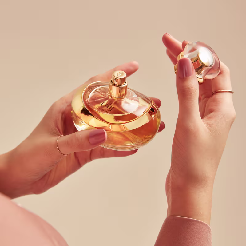 A imagem mostra um frasco de perfume Lily, feito de vidro transparente na cor âmbar. O frasco tem um formato arredondado elegante, com uma tampa dourada brilhante no topo. Uma mão está segurando o frasco.