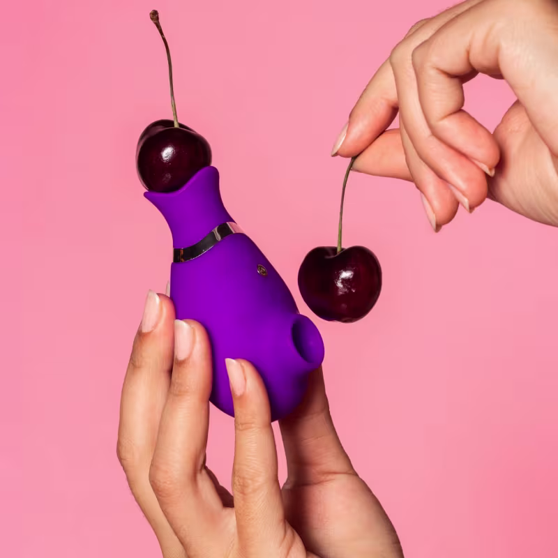 Vibrador roxo compacto com cereja vermelha, segurado em mãos femininas em fundo rosa.
