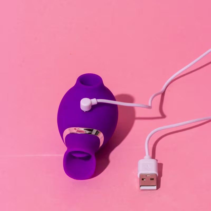 Vibrador recarregável de cor roxa, com cabo de carregamento USB conectado.