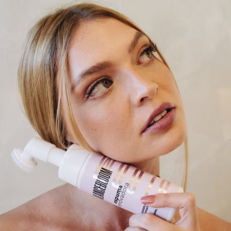 Mulher branca olhando diretamente para a câmera com um close em seu rosto. Ela tem cabelos loiros curtos e uma expressão serena. Ela está segurando o frasco de espuma de beleza da marca Bloom Cosméticos junto ao seu rosto.