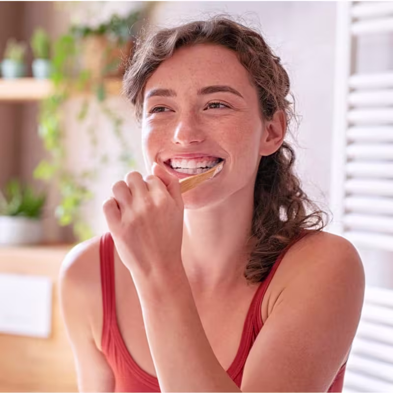 A imagem mostra uma jovem mulher sorridente aplicando um produto de higiene bucal com um aplicador descartável. Ela está usando um top vermelho de alças finas e tem cabelos castanhos ondulados.