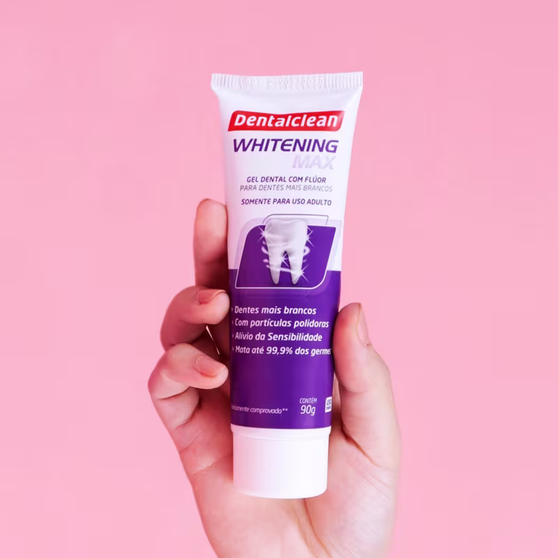 Imagem mostra o creme dental sendo segurado por uma mão branca com um fundo rosa atrás.