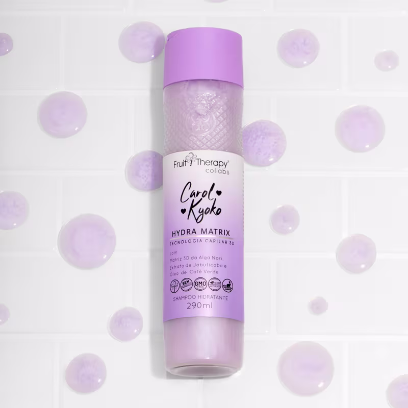 A imagem mostra o shampoo hidratante da linha Fruit Therapy Carol Kyoko. Ele tem um design cor lavanda e está posicionado no centro de uma superfície branca e gotas do líquido estão decorando o redor.