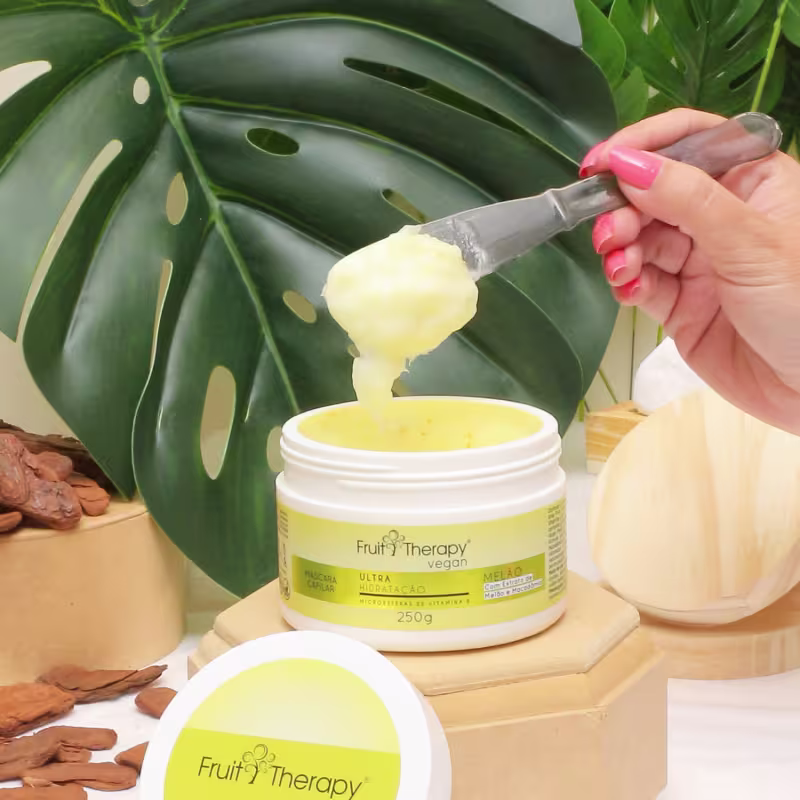 A imagem mostra a máscara de hidratação da linha Fruit Therapy Vegan de Melão. O pote branco com rótulo amarelo está aberto. Uma mão está retirando o conteúdo amarelado e cremoso com uma espátula. Ao redor, vemos folhas verdes tropicais.