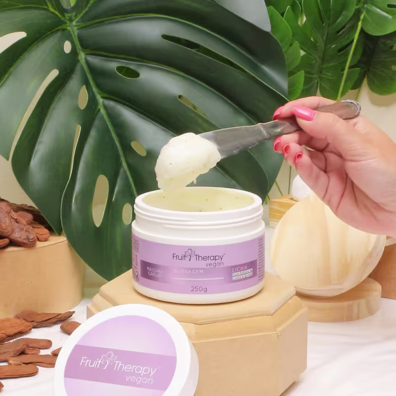 A imagem mostra a máscara Fruit Therapy Vegan Lichia Blindagem Pós-Química. O pote branco com rótulo rosa está aberto. Uma mão está retirando o conteúdo cremoso com uma espátula. Ao redor, vemos folhas verdes tropicais complementam a imagem.