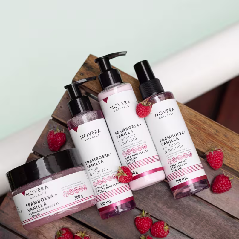 A imagem traz a linha Novera Naturals Framboesa + Vanilla. São 4 itens em embalagens de plástico verde e branca: esfoliante, sabonete líquido, loção corporal e body splash. Eles estão sobre uma superfície de madeira, com morangos espalhados ao redor.