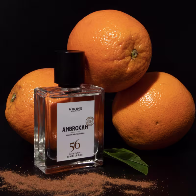 Perfume Viking Ambroxan em frasco retangular de vidro transparente com tampa preta. Rótulo branco com o nome do produto e número 56 em destaque. Três laranjas grandes ao redor do frasco, uma folha verde e especiarias espalhadas na superfície escura.