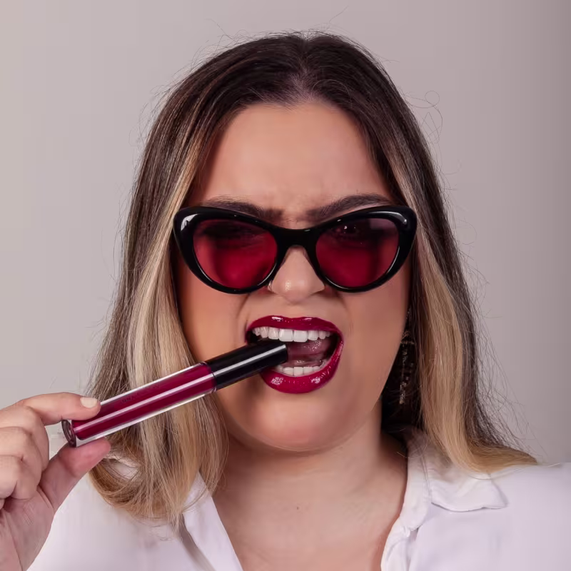 A imagem mostra uma mulher aplicando um batom líquido vermelho vivo em seus lábios. A mulher está usando óculos de sol grandes com lentes vermelhas e tem cabelos longos loiros e castanhos. Ela está sorrindo amplamente.