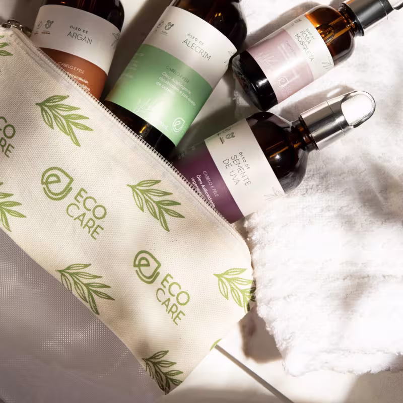 Frascos contendo óleos com argan e alecrim, com rótulos descrevendo os ingredientes naturais. Uma sacola de compras de tecido bege estampada com folhas estilizadas. Sobre um fundo branco, os produtos estão dispostos junto a uma toalha felpuda branca.
